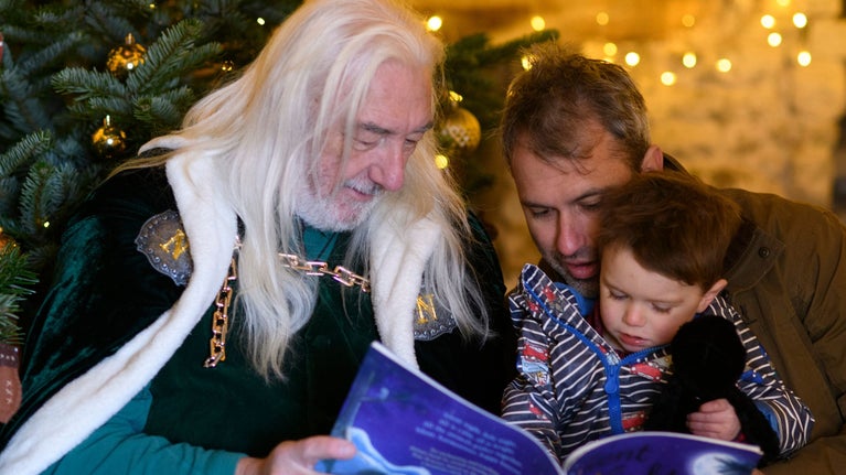 St Nicholas with long flowing white hair and a green cape reading a story to a boy sat on his father's lap in the Dovecote, surrounded by trees and glistening lights.